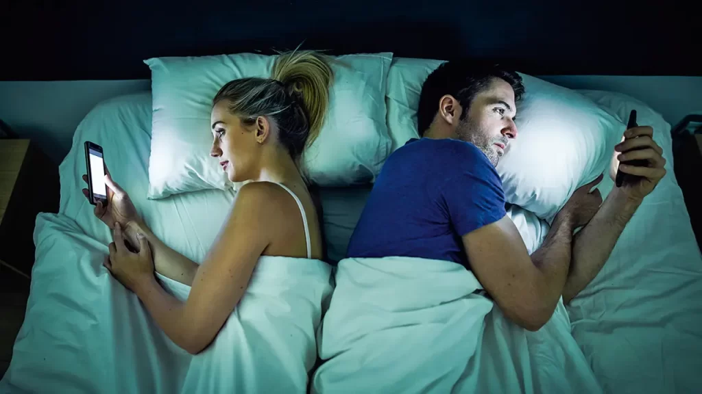 Casal deitado em uma cama, de costas um para o outro. Cada um segura seu celular na mão ignorando o parceiro.