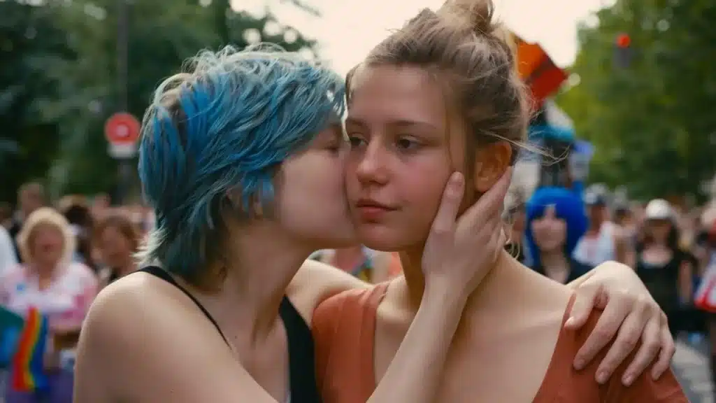 cena do filme azul é a cor mais quente, com o casal de garotas protagonistas do filme. A protagonista de cabelo azul está dando um beijo amoroso no rosto de sua parceira. 
