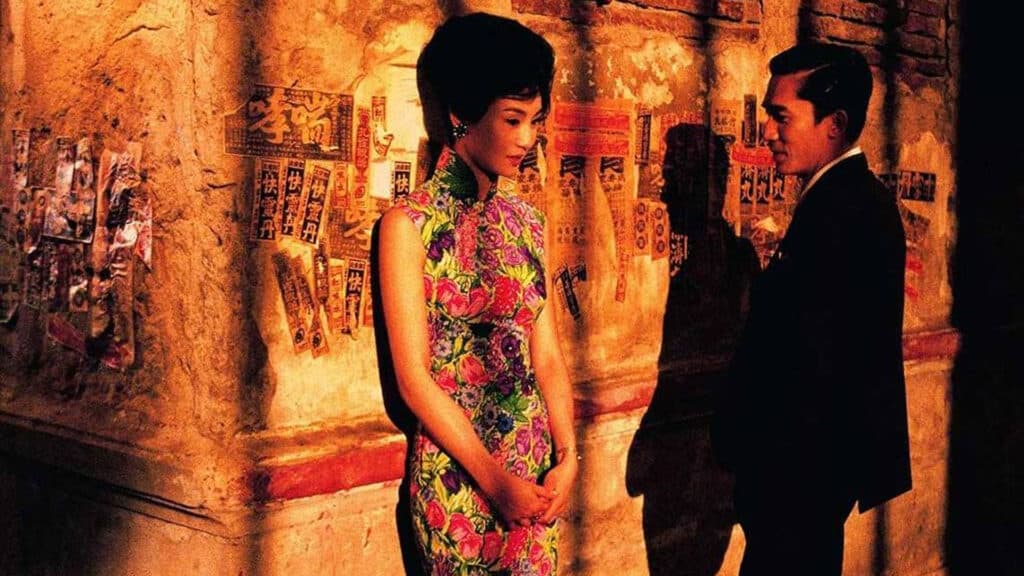 Casal asiático do filme Amor a flor da pele. A mulher está apoiada em uma prede na rua enquanto o homem se aproxima para falar com ela. Os dois mostram um sorriso no rosto. 